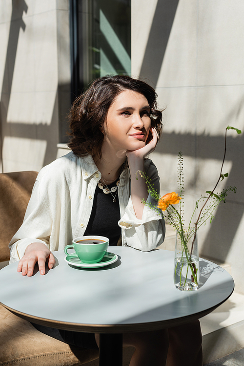 smiling woman sitting at white round table near cup of black aromatic coffee, saucer, glass vase with yellow rose and green plants on summer terrace of hotel cafe in morning sunlight