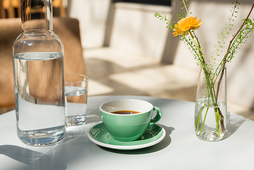 vase with yellow rose and green plants, glass and decanter with fresh pure water, saucer, cup with black coffee on white round table in morning sunlight, outdoor terrace of hotel cafe