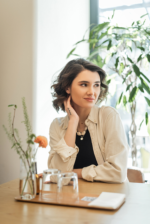 tattooed and carefree woman with wavy brunette hair looking away while sitting at table with glasses, napkins, vase with yellow rose and green plants on blurred foreground in hotel lobby cafe