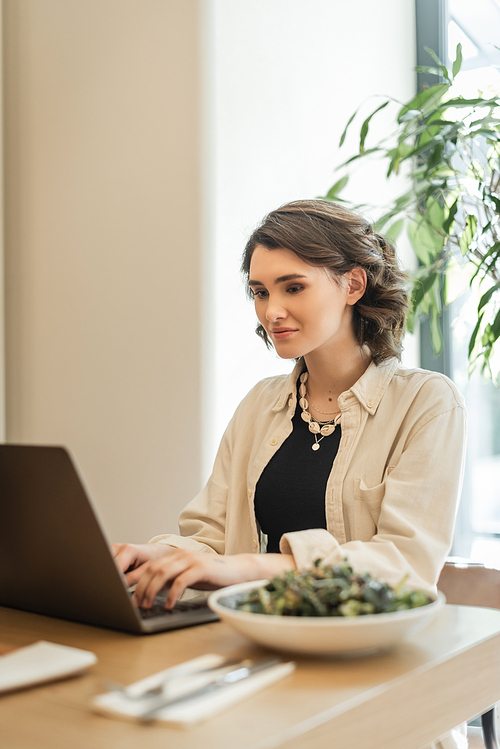 positive woman with wavy brunette hair, in stylish casual clothes typing on laptop near fresh vegetable salad while sitting at table in lobby cafe of contemporary hotel, work and travel