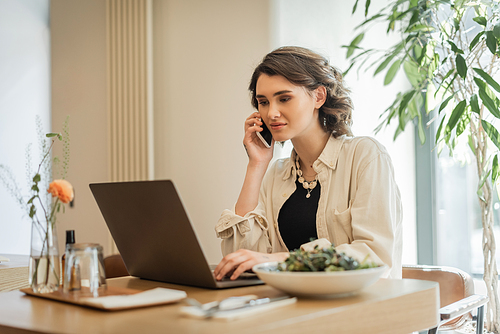 digital nomadism, young woman in stylish casual clothes, with wavy brunette hair talking on smartphone near laptop, vegetable salad, glass and vase with green plants in lobby cafe of modern hotel