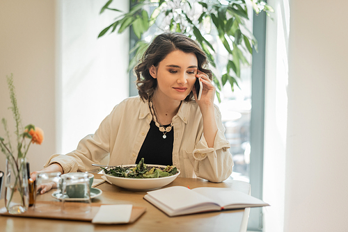 positive woman in stylish casual clothes looking at notebook during conversation on mobile phone near fresh vegetable salad, glasses and cup of aromatic coffee in hotel lobby, work-life integration