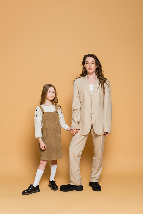 trendy mother and daughter, stylish working mother in suit holding hands with daughter while standing together on beige background, parent and child, fashionable family