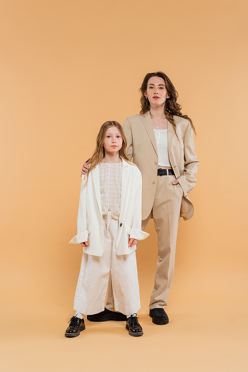 stylish mother and daughter in suits, woman and girl looking at camera while standing together on beige background, fashionable outfits, formal attire, corporate mom, modern family, hand in pocket