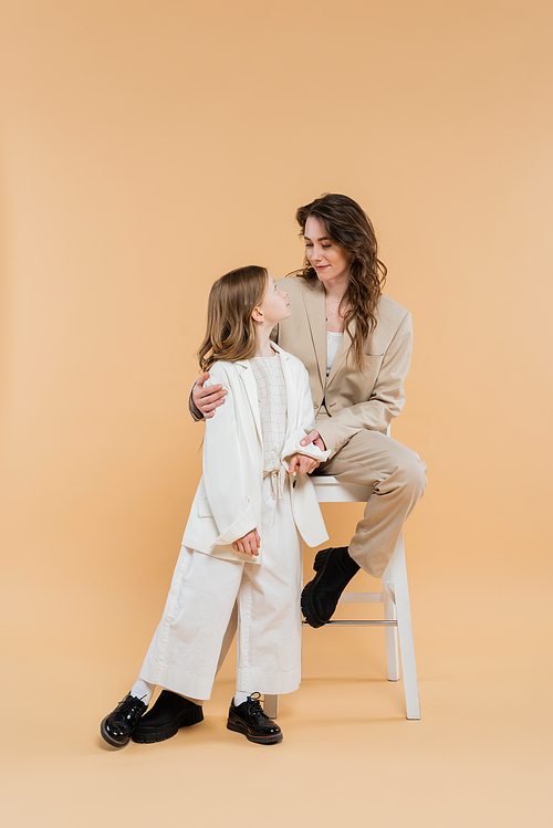 stylish mother and daughter in suits, woman sitting on high chair and looking at girl on beige background, fashionable outfits, formal attire, corporate mom, modern family