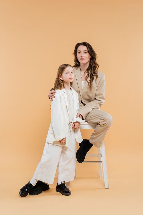 stylish mother and daughter in suits, woman and girl looking at camera, sitting on high chair on beige background, fashionable outfits, formal attire, corporate mom, modern family