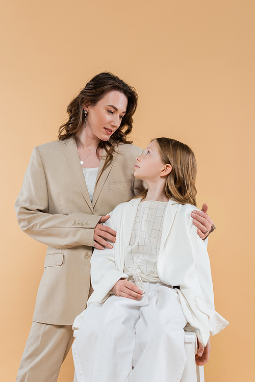 business mother and daughter in suits, woman hugging shoulders of girl sitting on chair on beige background, fashionable outfits, formal attire, corporate mom, modern family
