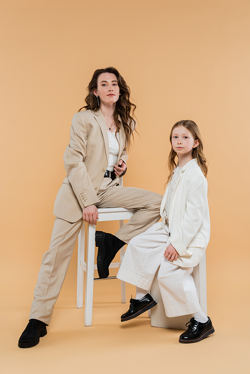 corporate mom and daughter in suits, woman sitting on high chair and looking at camera near girl on beige background, fashionable outfits, formal attire, corporate mom, motherhood and career