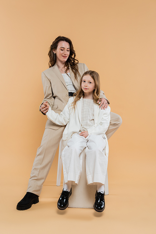 stylish mother and daughter in suits, woman and girl holding hands and sitting together on beige background, fashionable outfits, formal attire, corporate mom, modern family