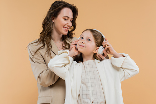 corporate mom and daughter in suits, happy woman wearing wireless headphones on girl while standing together on beige background, fashionable outfits, formal attire, modern family