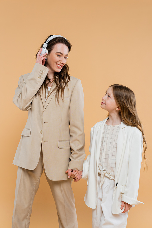happy mother and daughter in suits, woman wearing wireless headphones near girl and holding hands on beige background, fashionable outfits, formal attire, corporate mom, modern family