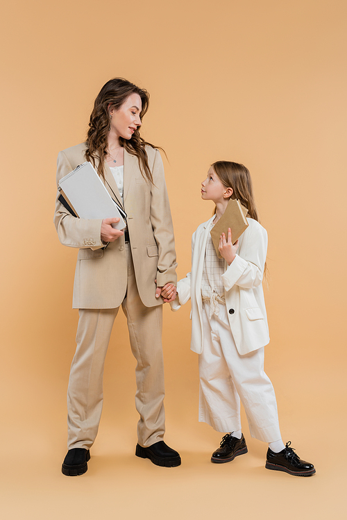 trendy mother and daughter in suits, woman and child holding books and notebooks while standing together on beige background, fashionable outfits, formal attire, corporate mom, education concept
