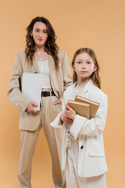 trendy mother and daughter in suits, child holding books near blurred woman with notebooks on beige background, fashionable outfits, formal attire, corporate mom, education concept