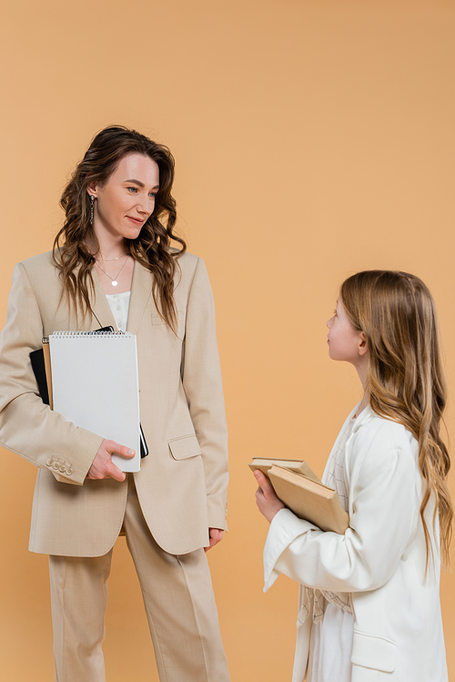 fashionable family, mother and daughter in suits, woman and child holding books and notebooks while standing together on beige background, formal attire, corporate mom, education concept