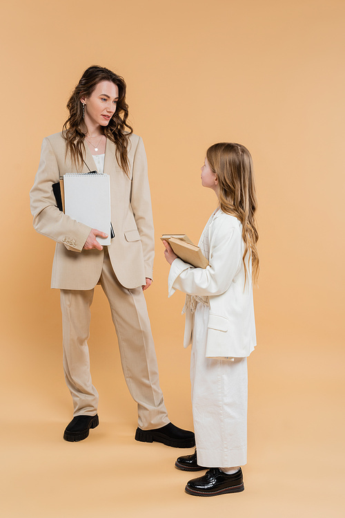 modern family, mother and daughter in suits, woman and child holding books and notebooks while standing together on beige background, formal attire, corporate mom, education concept