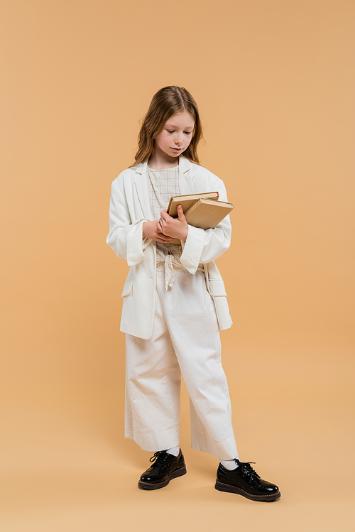 education concept, preteen girl in white suit holding books and standing on beige background, fashionable outfit, formal attire, back to school, preparing for school new year