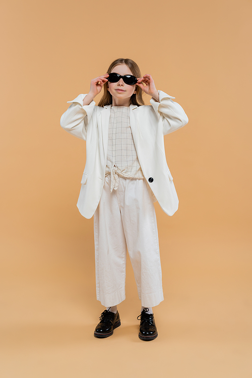 stylish preteen girl in white suit and black shoes looking at camera while wearing sunglasses and standing on beige background, fashionable outfit, formal attire, child model, trendsetter, style