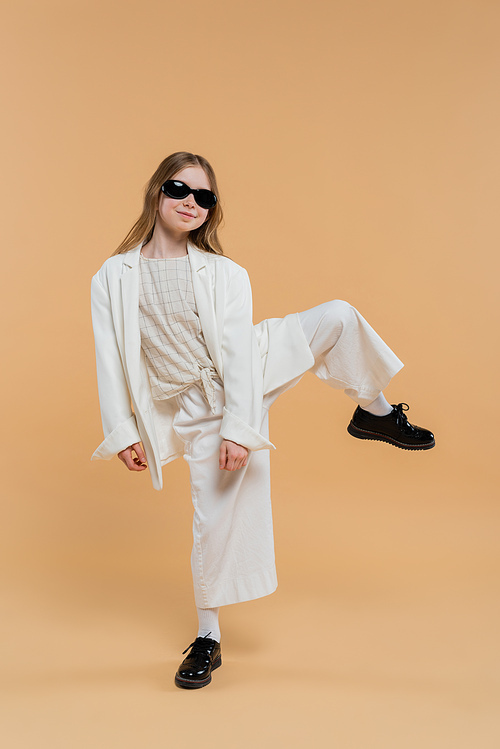 trendy preteen girl in white suit, sunglasses and black shoes posing with raised leg and standing on beige background, fashionable outfit, formal attire, child model, trendsetter, style