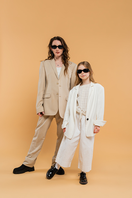 trendy mother and daughter in sunglasses, businesswoman and girl in suits  standing together on beige background, fashionable outfits, formal attire, corporate mom, modern family