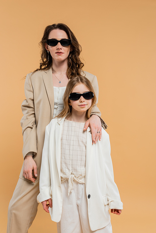 modern family, stylish mother and daughter in sunglasses, businesswoman and girl in suits standing together on beige background, fashionable outfits, formal attire, corporate mom