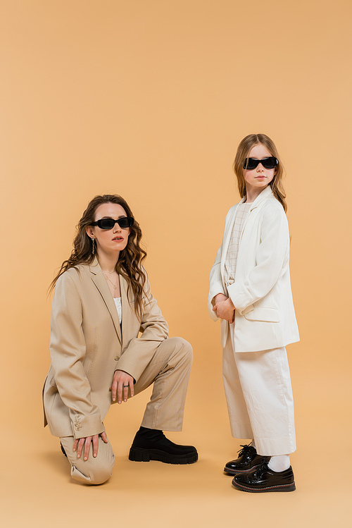 modern family, stylish mother and daughter in suits and sunglasses, businesswoman sitting near girl on beige background, fashionable outfits, formal attire, corporate mom