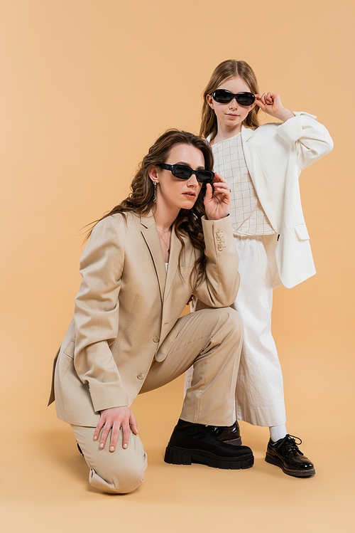 modern family, stylish mother and daughter in suits and sunglasses, businesswoman sitting near girl on beige background, fashionable outfits, formal attire, working mother, trendsetter