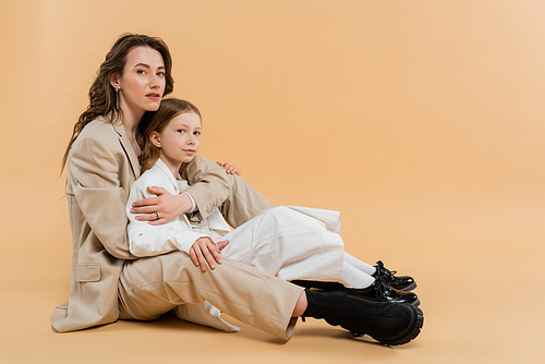 mother child bonding concept, stylish woman in suit hugging preteen daughter and standing together in suits on beige background, corporate mom, businesswoman, motherly love
