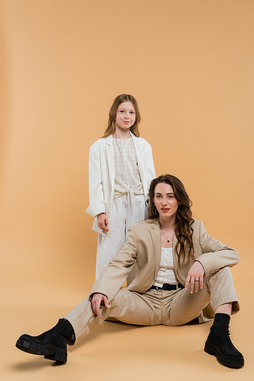 corporate mom concept, stylish woman in suit sitting near preteen daughter and looking at camera on beige background, togetherness businesswoman, motherly love