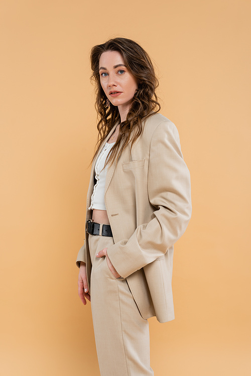 style and fashion concept, young woman with wavy hair standing in fashionable suit while posing on beige background, formal attire, hand in pocket, looking at camera, modern elegance