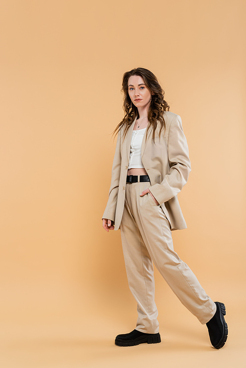fashion trend concept, young woman with wavy hair walking in fashionable suit and looking at camera on beige background, hand in pocket, classic style, professional attire