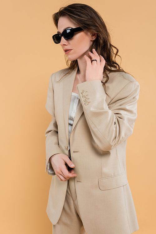 fashion trend concept, young woman with wavy hair standing in fashionable suit and sunglasses on beige background, classic style, chic stylish posing, professional attire, formal attire