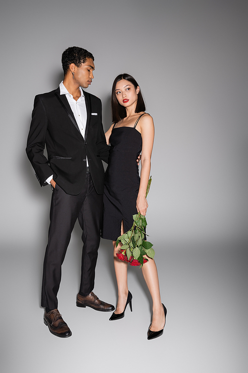 full length of african amrican man in black stylish suit embracing asian woman holding red roses on grey background