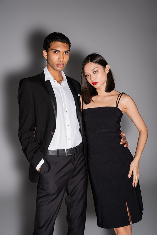 african american man in black suit and asian woman in strap dress looking at camera on grey background