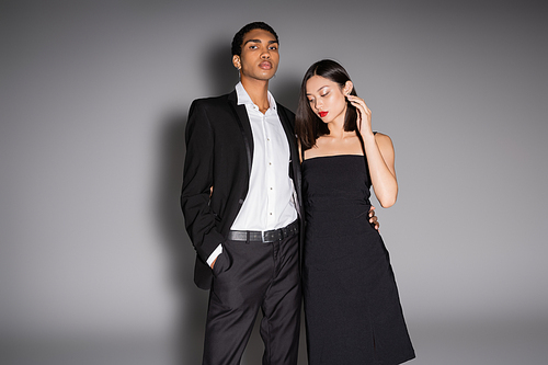 african american man in elegant suit looking at camera near asian woman in black dress on grey background