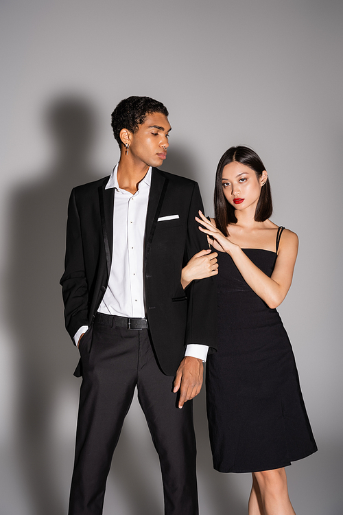 brunette asian woman in black dress looking at camera near elegant african american man standing with hand in pocket on grey background