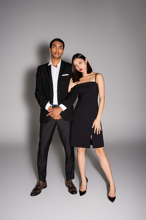 full length of interracial couple in elegant formal wear looking at camera on grey background