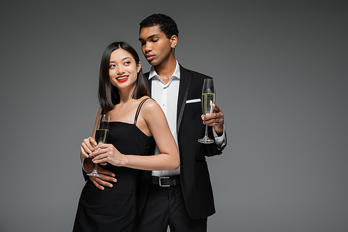 smiling asian woman and elegant african american man holding champagne glasses isolated on grey