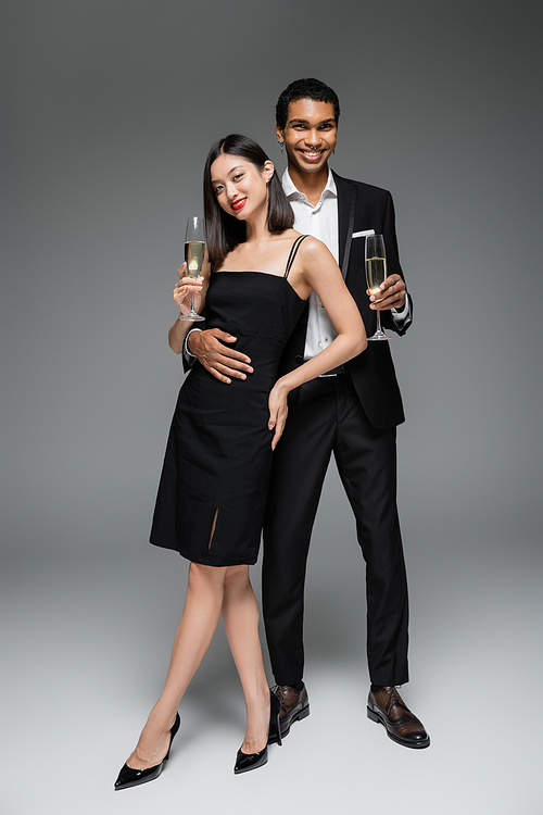 full length of cheerful african american man embracing elegant asian woman while standing with champagne glasses on grey background