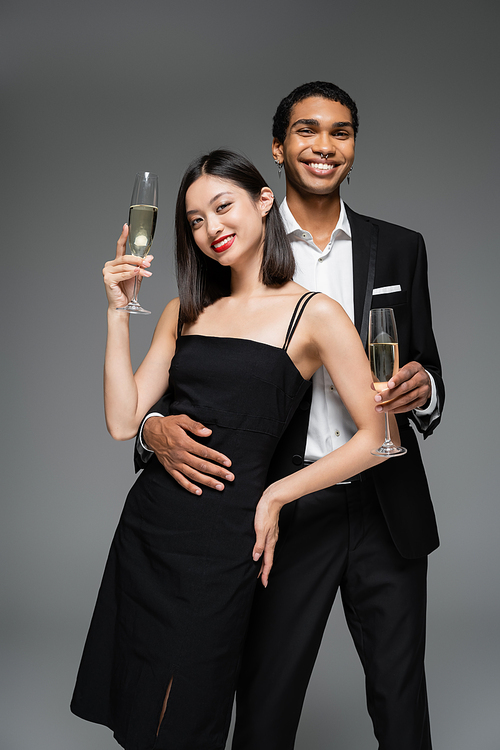 interracial couple in elegant attire holding champagne glasses and smiling at camera isolated on grey