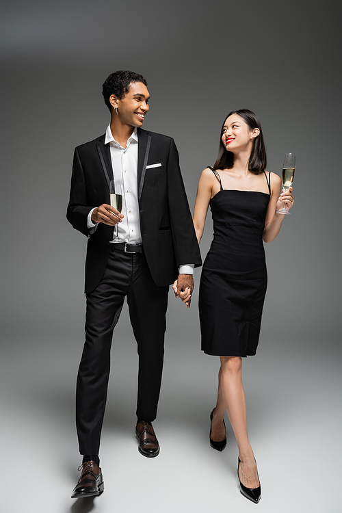 full length of elegant multiethnic couple with champagne glasses holding hands and smiling at each other while walking on grey background