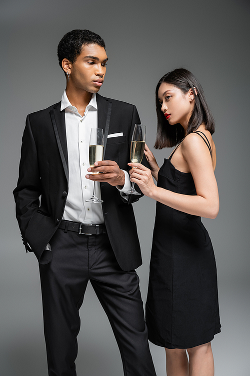 elegant african american man in suit and asian woman in black dress posing with champagne glasses isolated on grey