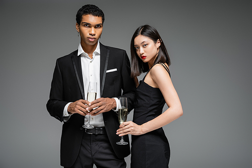 african american man in black suit and asian woman in strap dress holding champagne and looking at camera isolated on grey