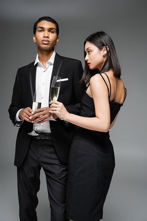 young interracial couple in black elegant clothes posing with champagne glasses isolated on grey