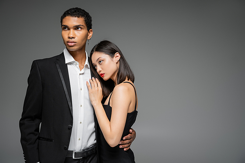 sensual asian woman in strap dress leaning of elegant african american man in black blazer isolated on grey