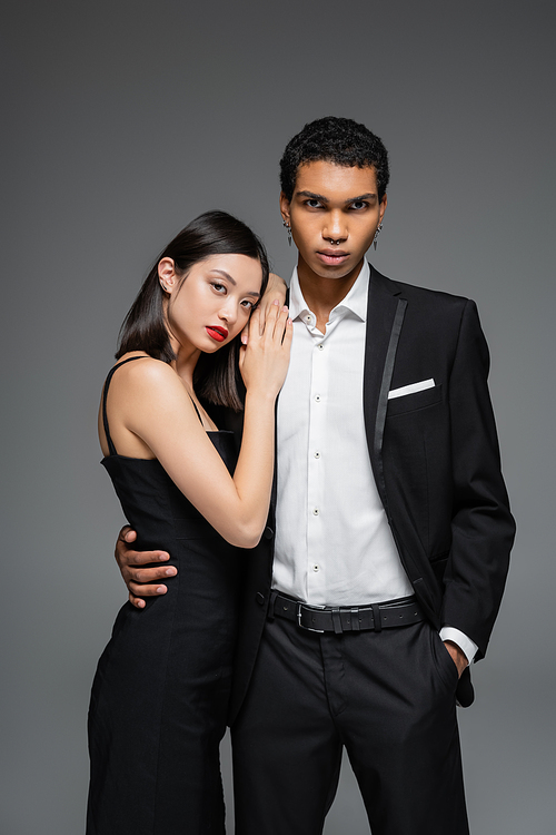 elegant african american man standing with hand in pocket and hugging sensual asian woman with red lips isolated on grey