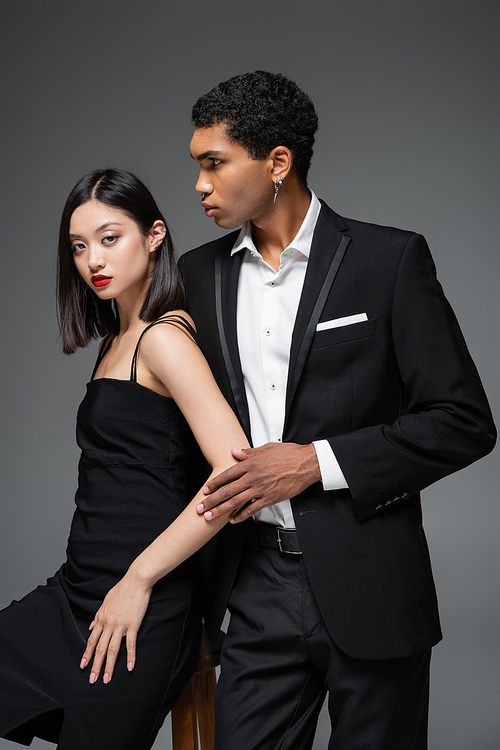 african american man in black blazer looking away near asian woman in elegant strap dress isolated on grey