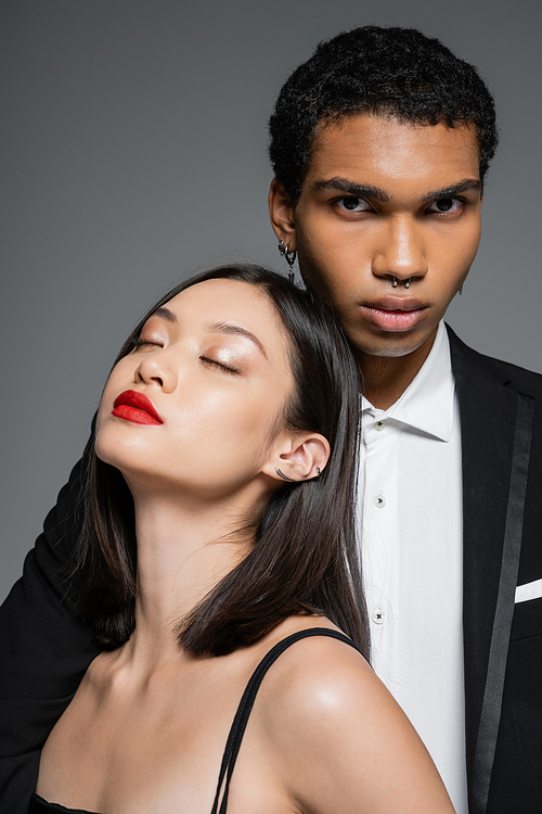 stylish african american man looking at camera near sensual asian woman with red lips and closed eyes isolated on grey