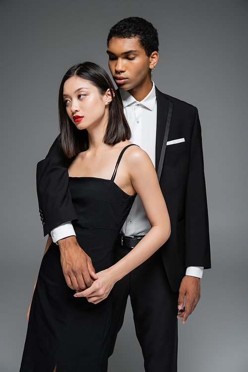 fashionable african american man embracing asian woman in black elegant suit looking away isolated on grey