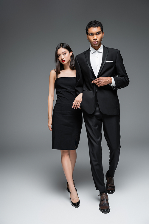 full length of multiethnic couple in elegant formal wear walking and looking at camera on grey background