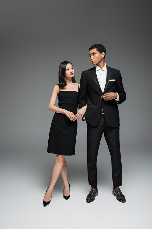 full length of interracial couple in black elegant attire looking at each other on grey background